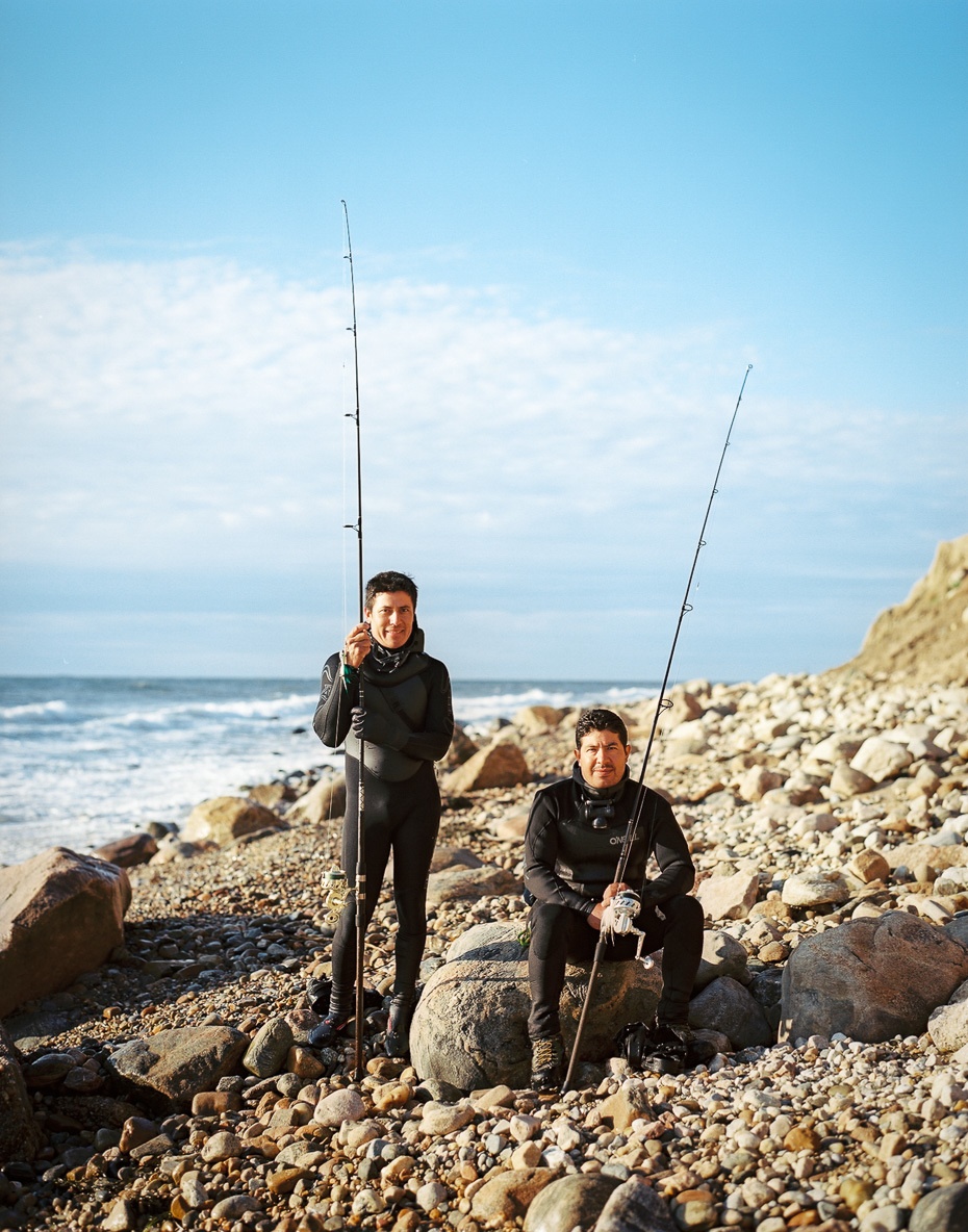 2 men in wetsuits holding fishing rods at the beach