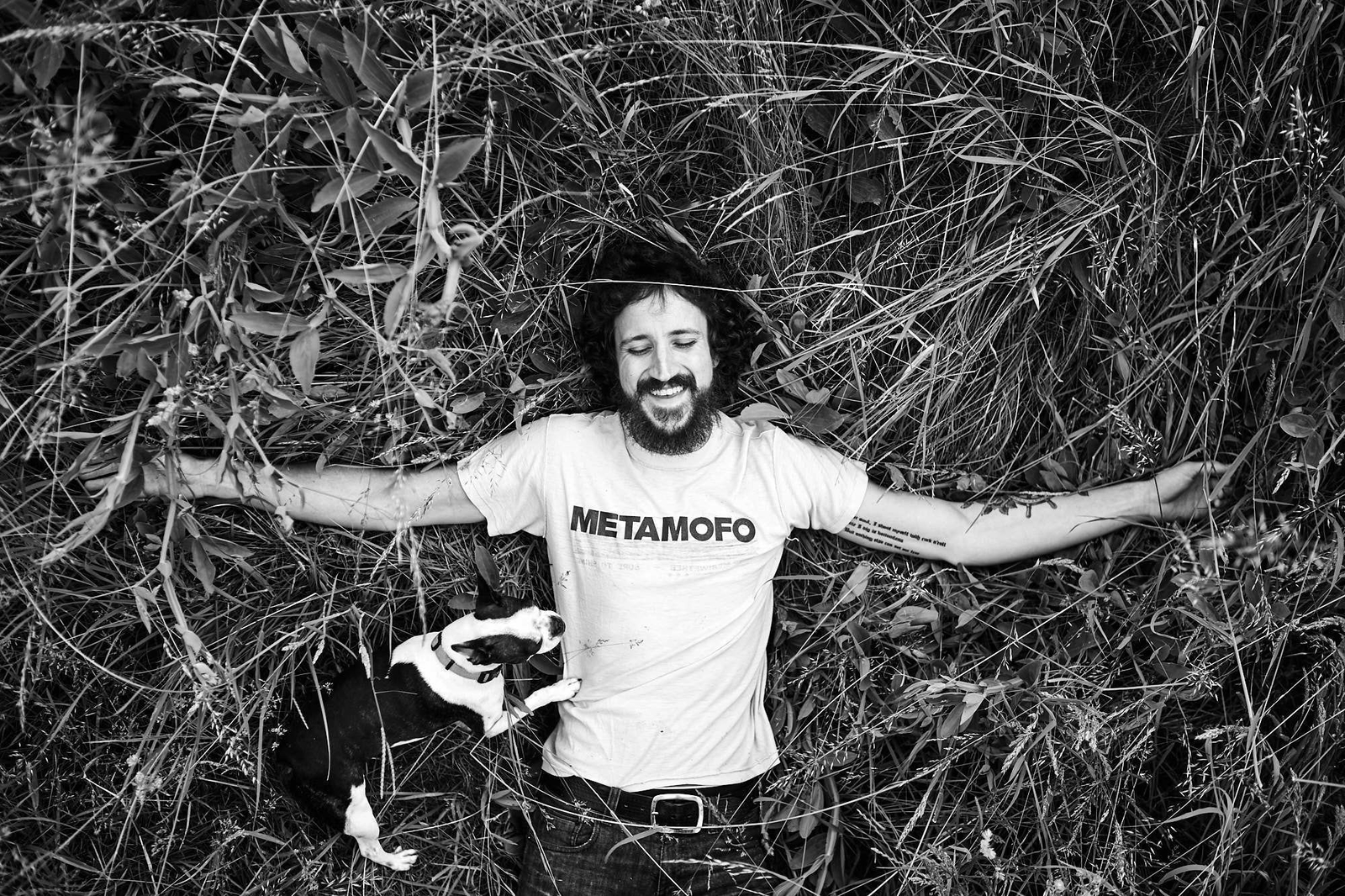 Laughing man lies in grass with black and white dog and Metamofo shirt