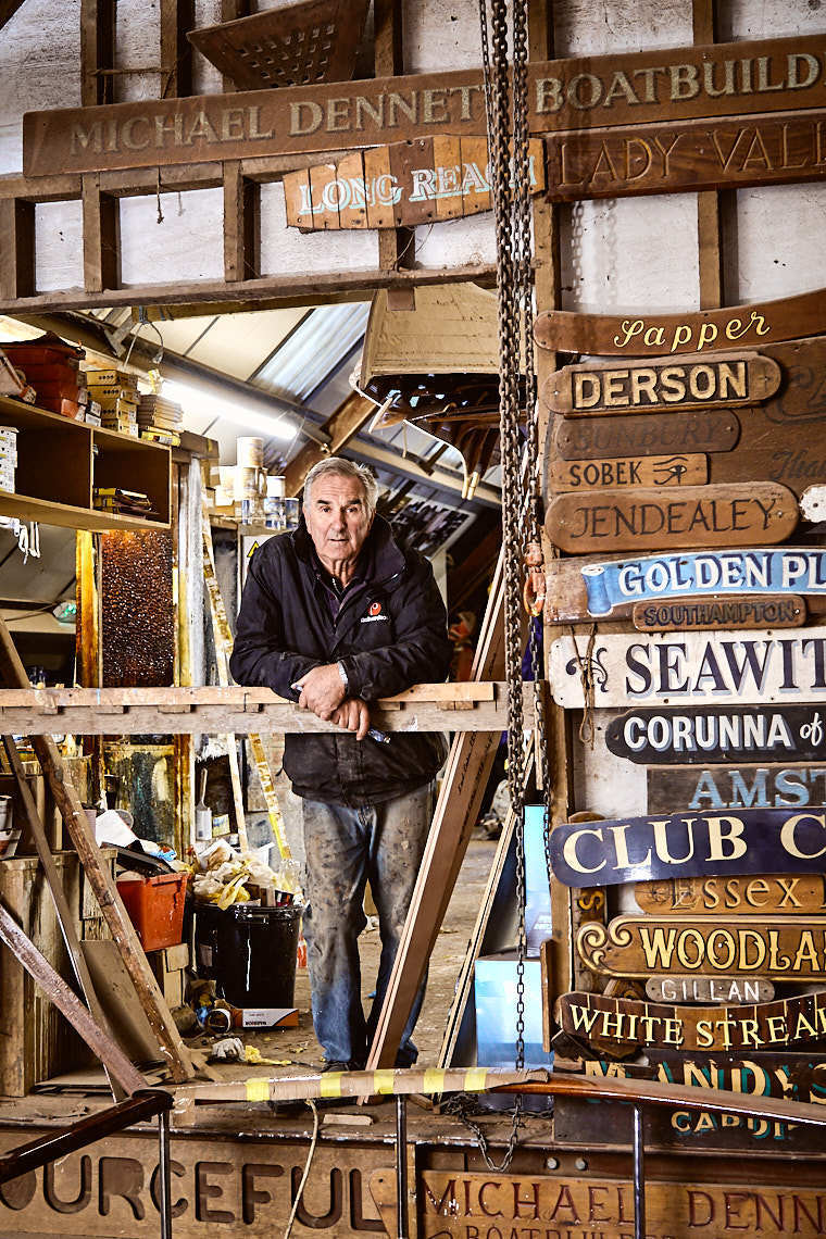 Michael Dennett Boat Builder man stands in shop with wooden signs
